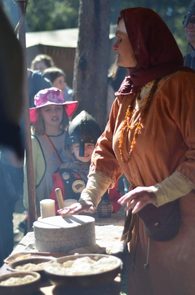 Children watching a woman grind grain by hand is a scene that has happened a thousand years in a row. You are not photographing history — you are photographing continuity.