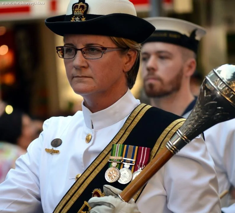 The white dress uniform, the ceremonial mace, the row of medals — everything about the image says service. The posture says the rest.
