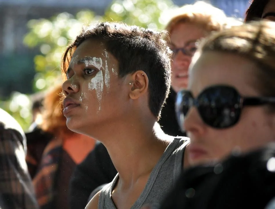 White ceremonial paint on a face in a crowd. The two registers — ancient and present — exist simultaneously without contradiction.