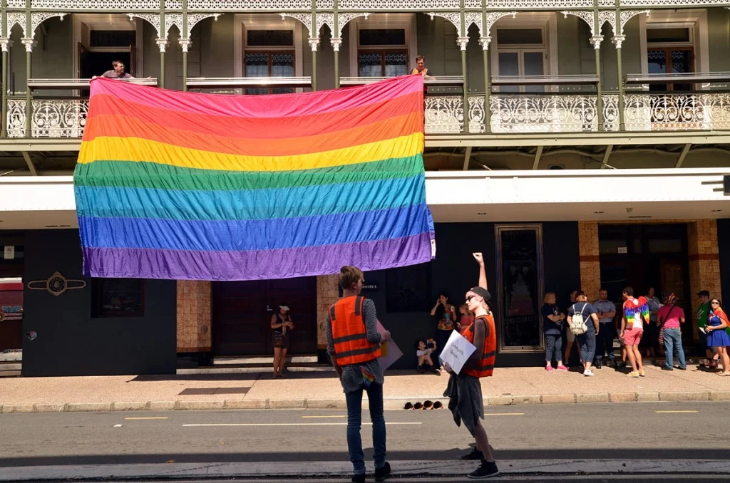 A full-width rainbow flag draped from an iron-lace balcony while a marshal fist-pumps below. The building has been there for over a century. So has the fight.