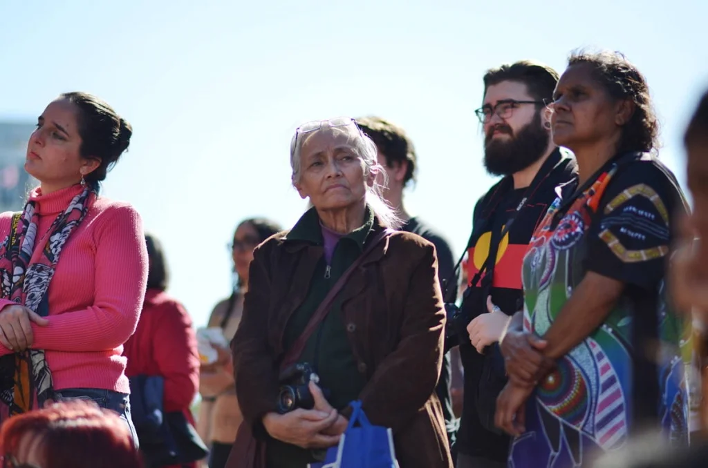 An elder in a brown coat holding a camera, listening with the stillness of someone who has been at this for decades. Some of the best photographs at a rally are of people watching.