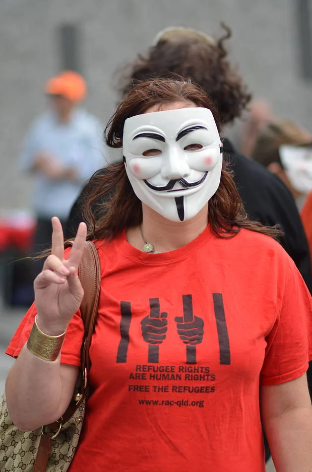 Guy Fawkes mask, red refugee rights shirt, peace sign. The image makes its argument without requiring a caption.