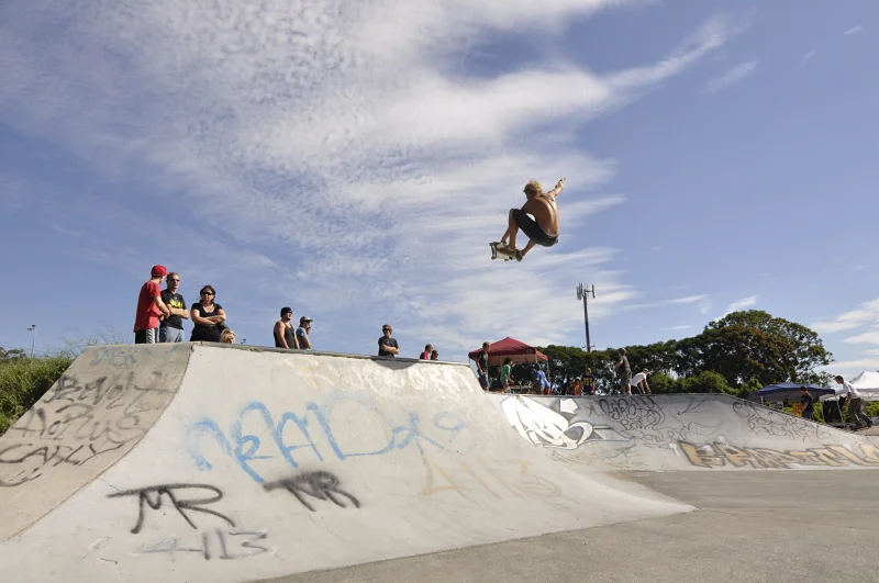Wide shot of the skate park, the skater at the top of his arc above the half-pipe. The crowd and the venue are all visible. The context makes the height legible.