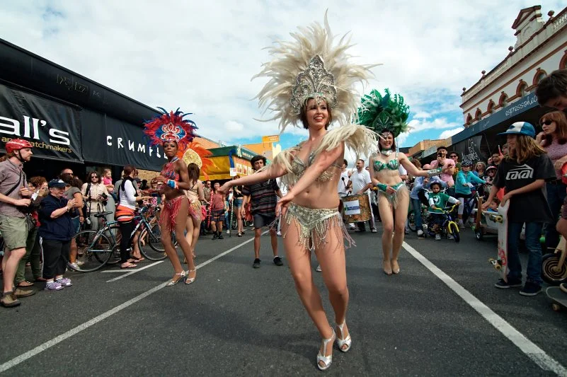 Samba dancers coming up Boundary Street, white feathers above the crowd, the heritage shopfronts behind them. West End contains multitudes.