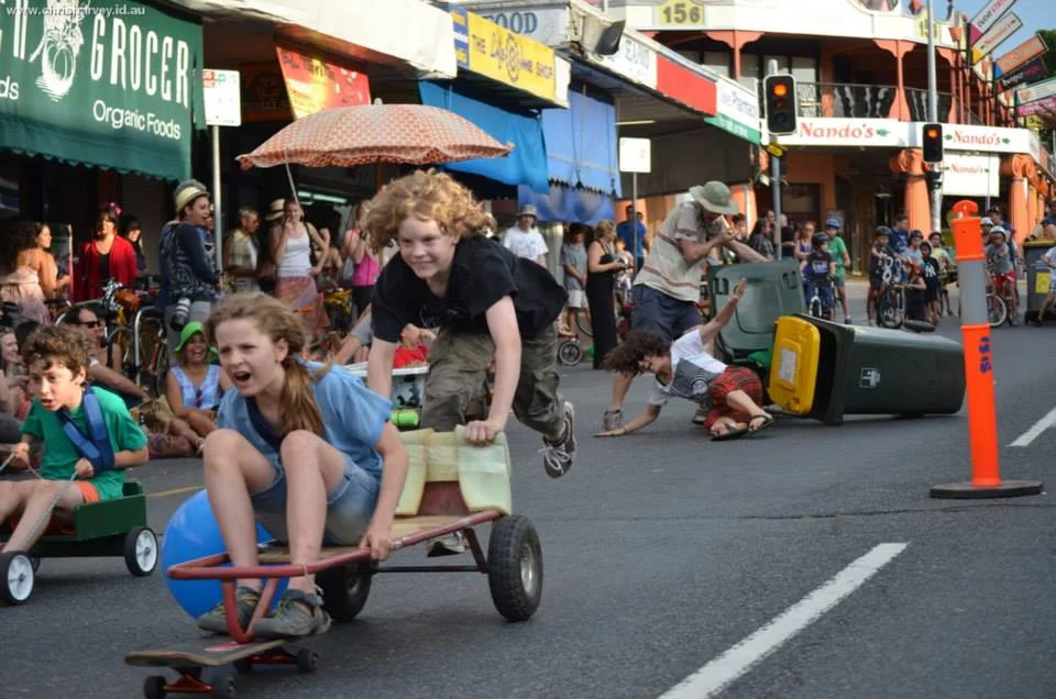 A go-kart race at dusk on Boundary Street — the girl at the front laughing, the boy behind pushing hard, the whole street behind them in chaos.