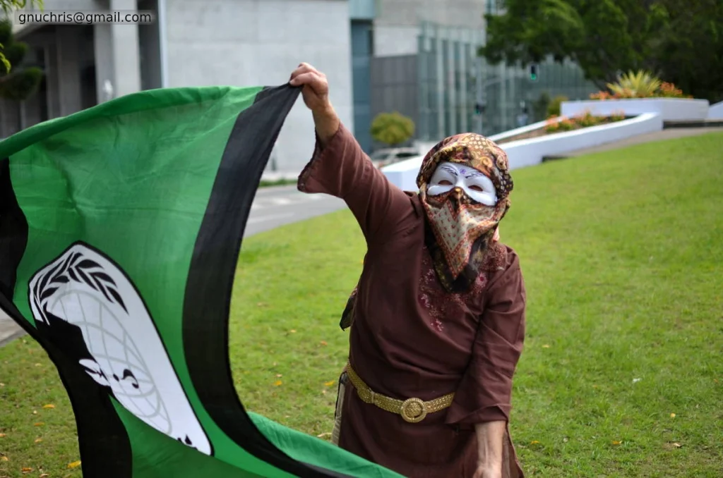The Anonymous flag in one hand, an ornate mask over the face, the whole costume assembled from different registers. She has put more thought into this than most.