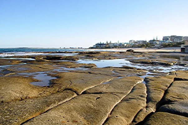 The wide view — rock shelf, sand, town, sky. It takes a minute to stop moving and see the frame that was there all along.