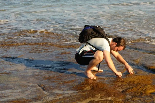 Down low on the rocks, backpack on, looking into the pool. The coast rewards the people who get off the path.