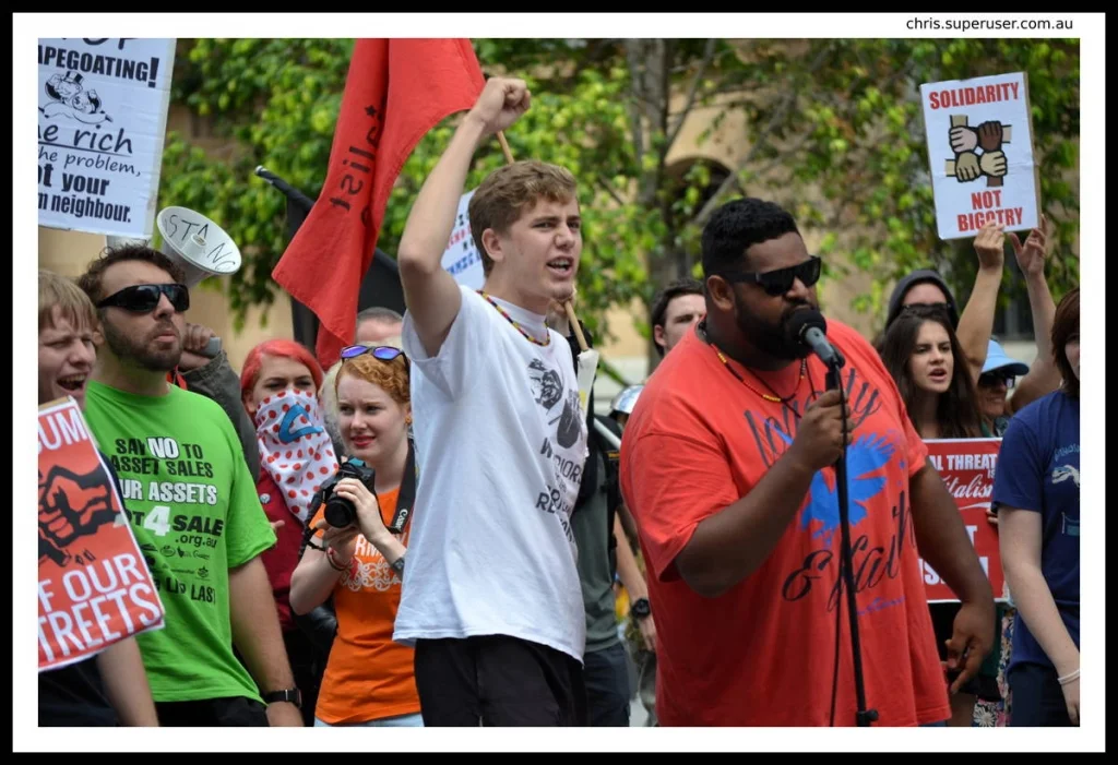 The person speaking at a rally microphone is not performing. They are arguing. The difference shows in the body — the lean forward, the fist, the urgency in the jaw.